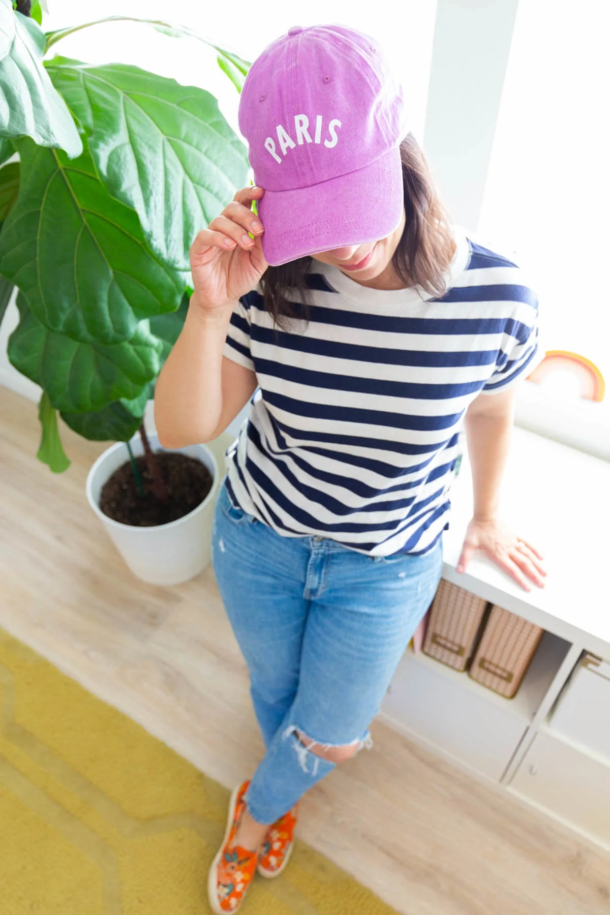 Woman in jeans and striped tshirt wearing a pink baseball cap with the word paris.