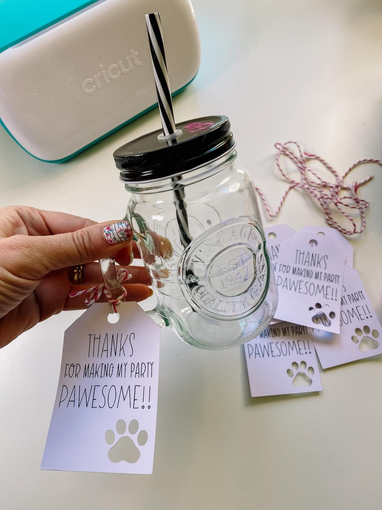 Hand holding a glass drink jar with black lid with a gift tag, with visible gift tags and cricut machine in background.