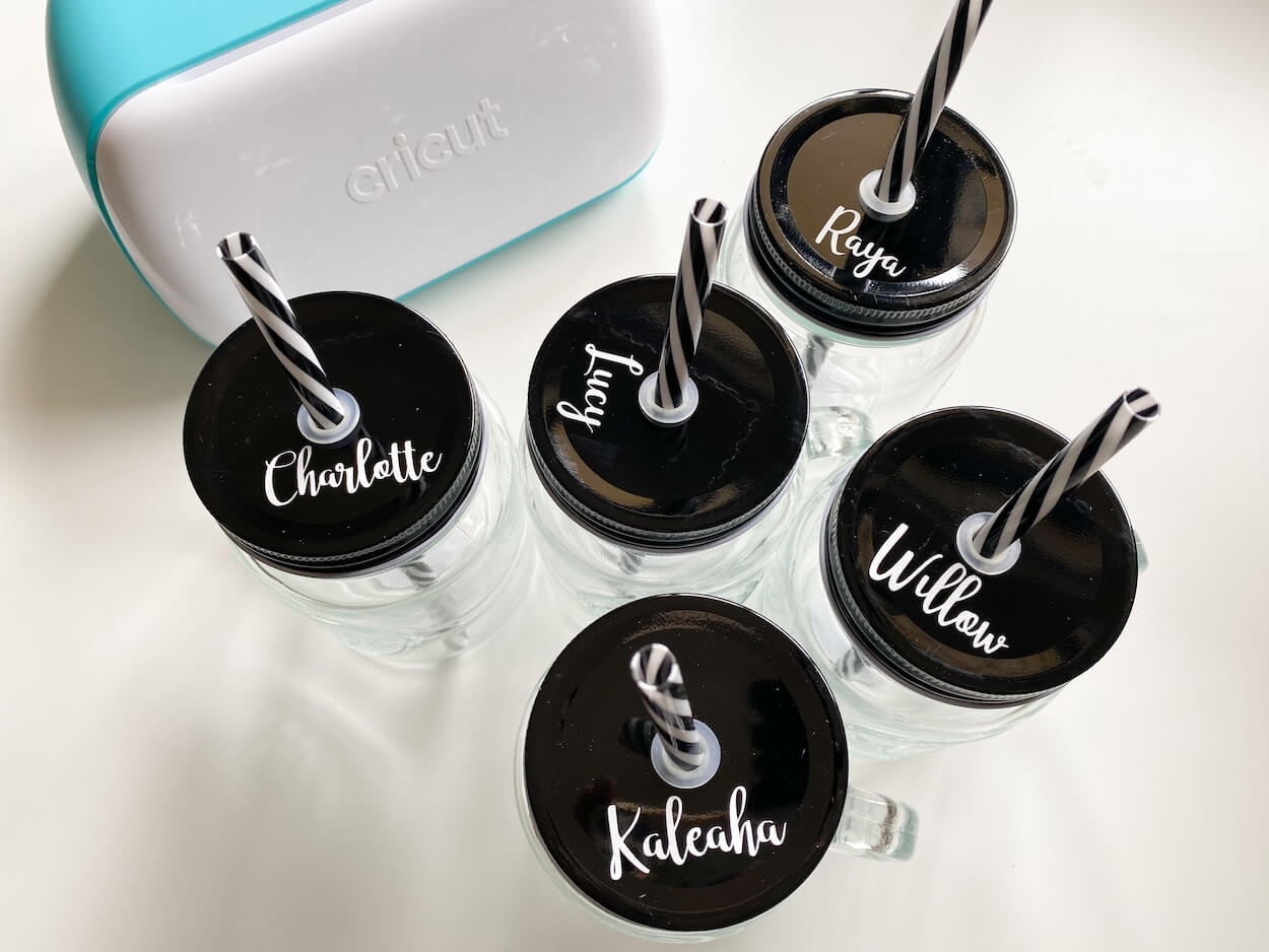 Drink jars with black lids personalised with names, next to a cricut joy machine.